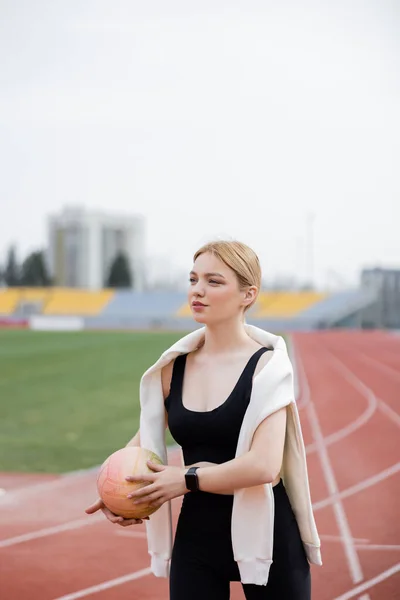 Молода спортсменка, дивлячись далеко, тримає м'яч на стадіоні — стокове фото