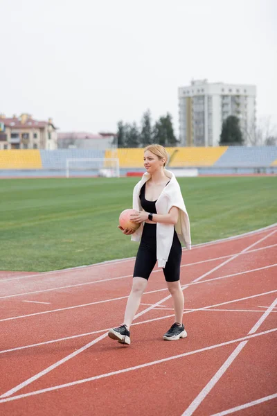Full length view of smiling sportswoman walking on stadium with ball — Stock Photo