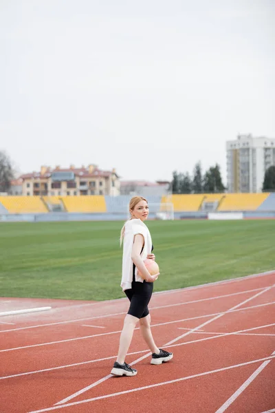 Full length view of young sportswoman looking at camera while walking with ball on stadium — Stock Photo