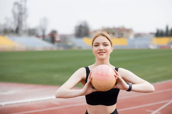 Cheerful sportswoman looking at camera while training with ball outdoors — Stock Photo