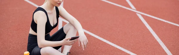 Cropped view of sportswoman sitting on athletic field with mobile phone, banner — Stock Photo