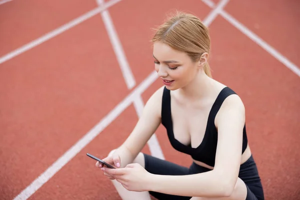 Усміхнена спортсменка використовує мобільний телефон, сидячи на стадіоні — стокове фото