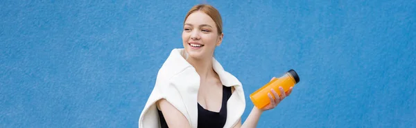 Cheerful sportive woman with orange juice looking away on blue textured background, banner — Stock Photo