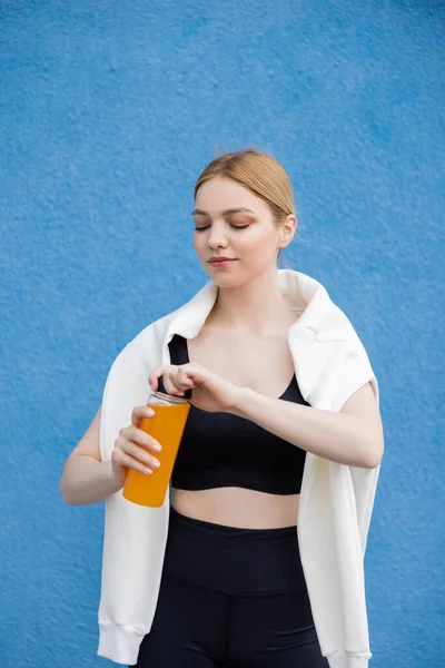 Sportive woman in black sports bra opening bottle of fresh orange juice on blue textured background — стоковое фото