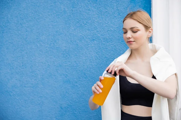 Mujer joven y deportiva que abre la botella de jugo de naranja cerca de la pared de textura azul - foto de stock