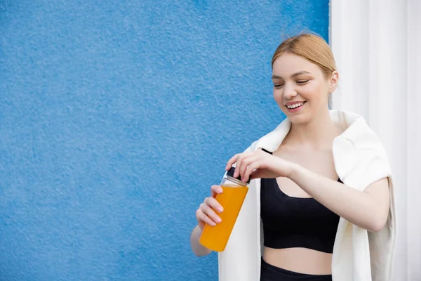 Donna sorridente in reggiseno sportivo nero contenitore di apertura con succo d'arancia vicino alla parete blu — Foto stock