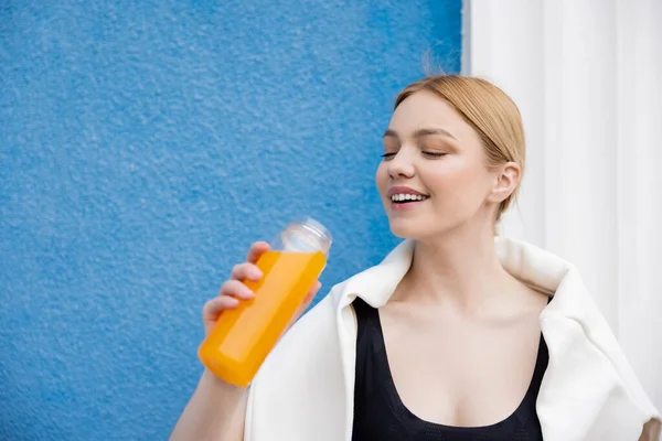 Sportswoman soddisfatto che tiene la bottiglia di succo d'arancia rinfrescante vicino alla parete blu — Foto stock