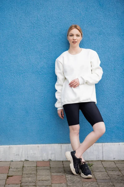 Full length view of woman in white sweatshirt and black bike shorts near blue wall outdoors — Stock Photo