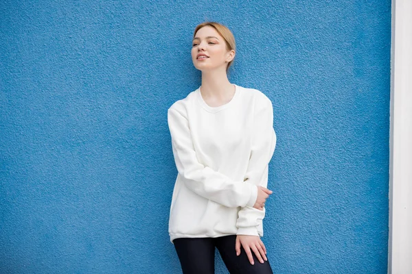Блондинка в білому светрі, дивлячись далеко біля синьої текстурованої стіни — стокове фото