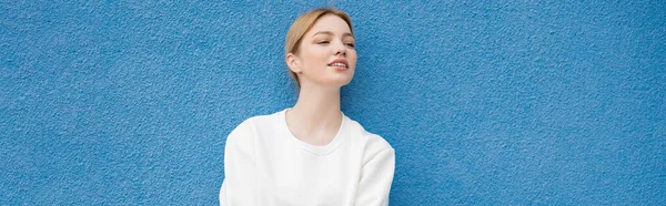 Lächelnde Frau in weißem Sweatshirt, die in der Nähe der blauen Wand wegschaut, Banner — Stockfoto