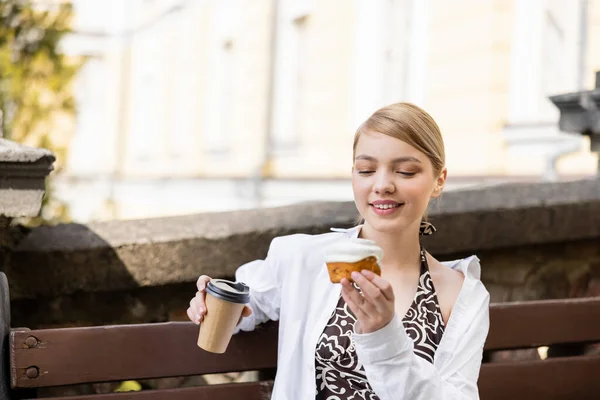 Jolie et heureuse femme assise sur un banc avec cupcake et café à emporter — Photo de stock