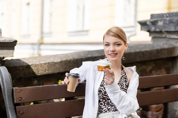 Mujer bonita con café para ir y cupcake sentado en el banco y mirando a la cámara — Stock Photo