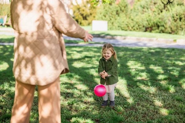 Petite fille en vêtements à la mode jouant avec la balle sur la pelouse verte près de mère floue — Photo de stock