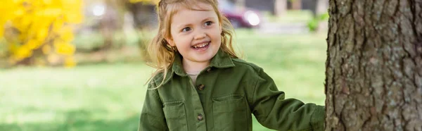 Happy girl looking away while standing near tree in park, banner — Stock Photo