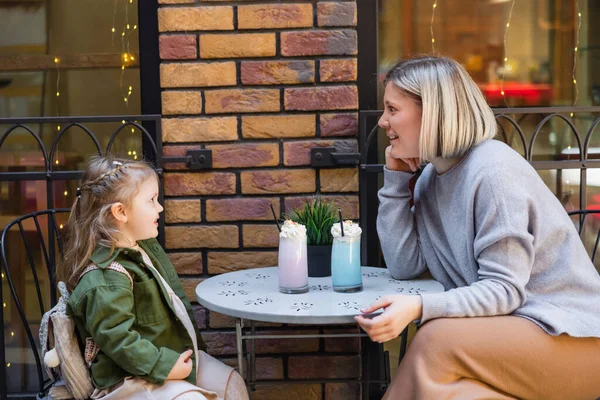 Seitenansicht einer aufgeregten Frau, die neben Tochter und Gläsern mit Milchshake im Straßencafé sitzt — Stockfoto