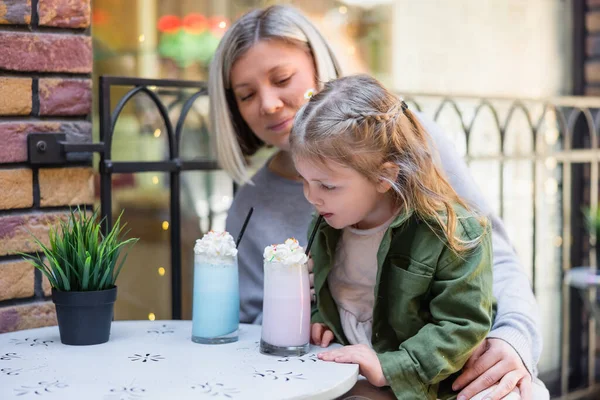 Kleines Mädchen trinkt süßen Milchshake in der Nähe von Mutter und Blumentopf in Café im Freien — Stockfoto