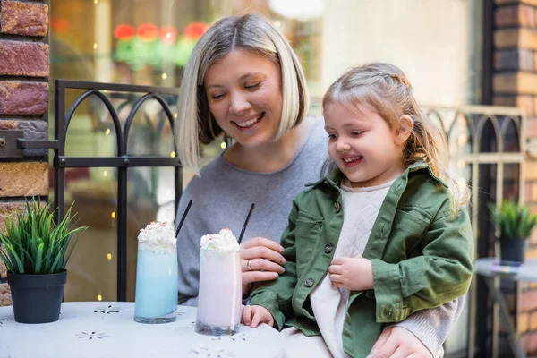 Lächelndes Mädchen mit Blick auf köstliches Milchdessert in der Nähe einer glücklichen Mutter im Straßencafé — Stockfoto