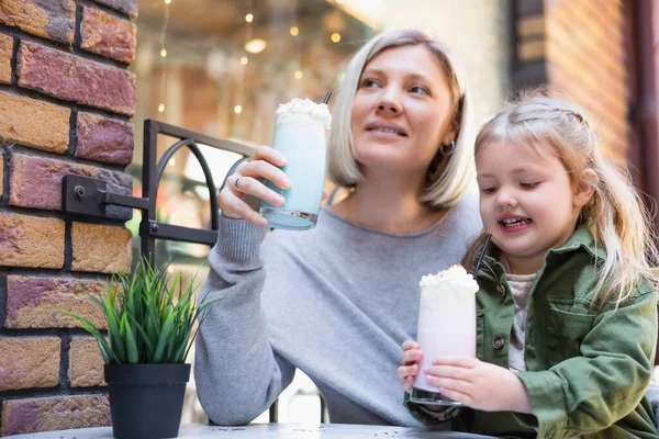 Niña sonriente sosteniendo un vaso de batido cremoso cerca de la pequeña hija en la cafetería de la calle - foto de stock