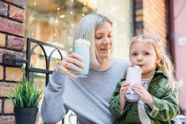 Kleines Mädchen trinkt leckeren Milchshake neben lächelnder Mutter in Straßencafé — Stockfoto