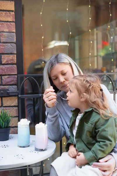 Happy woman feeding daughter with milk dessert in cafe on urban street — Stock Photo