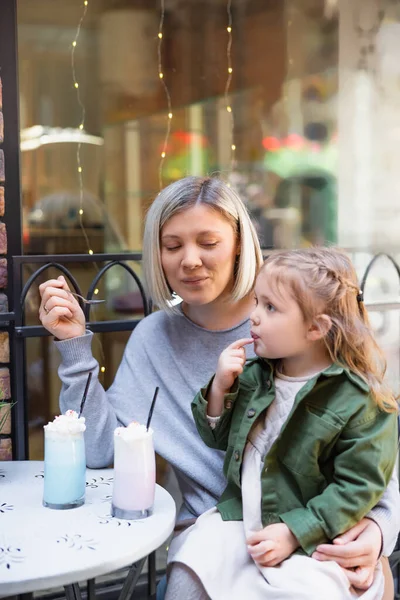 Smiling woman holding spoon near daughter and tasty milkshake in street cafe — Stockfoto