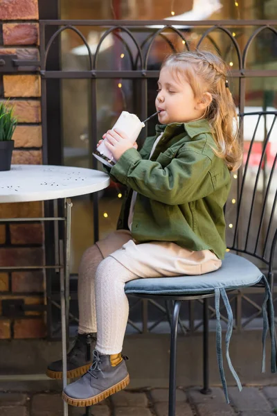 Full length view of girl sitting on chair in street cafe and drinking milkshake — Stock Photo