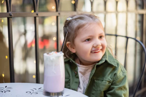 Cheerful child looking away near blurred glass of milkshake outdoors — Stock Photo