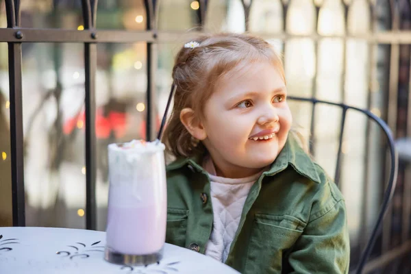 Chica feliz mirando lejos cerca borrosa vaso de batido en la cafetería de la calle - foto de stock