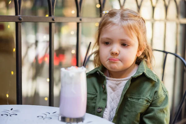Girl pouting lips while looking at blurred milkshake in street cafe — Stock Photo