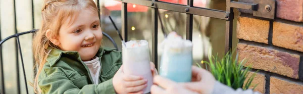 Souriant fille toasting avec des lunettes de milkshake près de maman floue à l'extérieur, bannière — Photo de stock