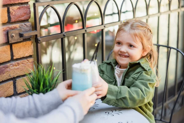 Cheerful girl clinking glasses with tasty milkshake with blurred mom in cafe outdoors — стоковое фото