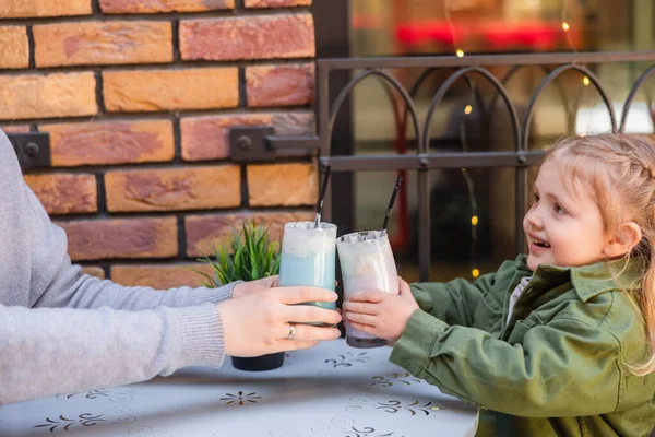 Happy girl toasting with glasses of milkshake while sitting with mother in street cafe — Stock Photo