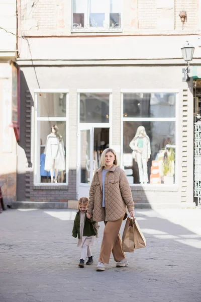 Madre con bolsas de compras caminando con hija alegre en la calle urbana - foto de stock