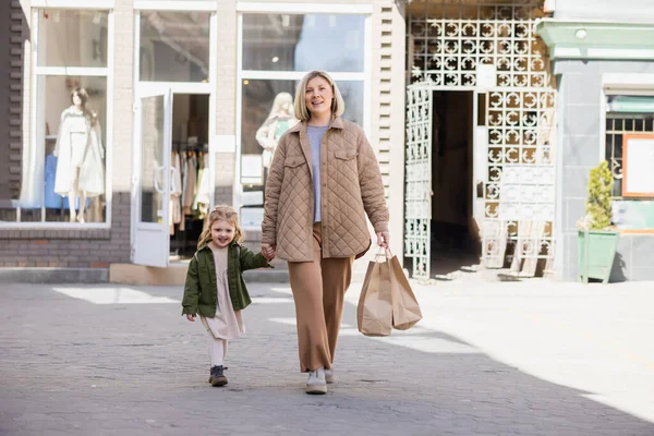 Donna sorridente con borse della spesa guardando la macchina fotografica mentre cammina con il bambino per strada — Foto stock