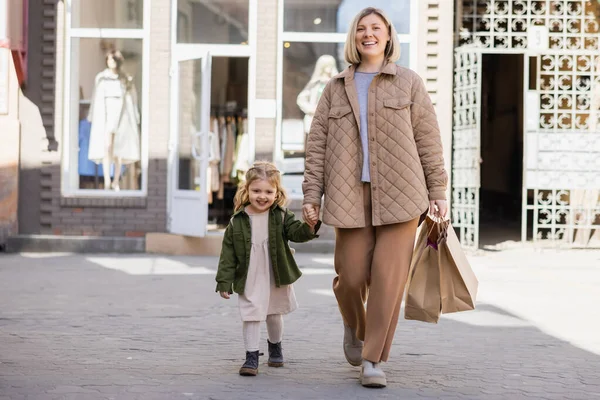 Cheerful mother and daughter in trendy clothes walking with shopping bags in city — Photo de stock