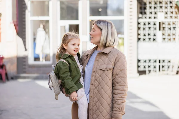 Blonde woman in trendy clothes holding smiling child on city street — Foto stock