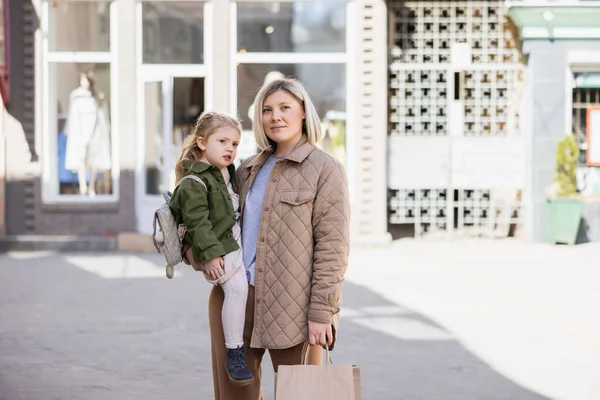 Positive and trendy woman with shopping bags looking away while holding daughter outdoors — стоковое фото