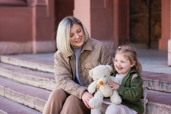 Trendy and happy mother and child sitting on city street with teddy bear — Stock Photo