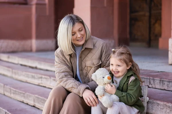 Joyful child sitting on stairs with mother and teddy bear — Stock Photo