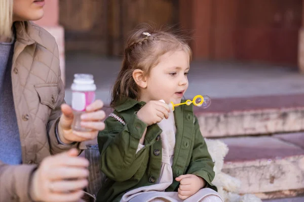 Child blowing soap bubbles near blurred mother outdoors — стоковое фото