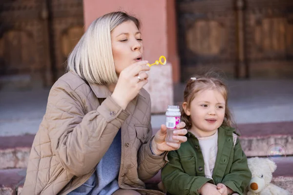 Stylish woman blowing soap bubbles near smiling daughter on urban street — Stock Photo