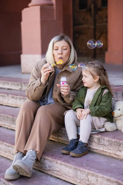 Blonde woman blowing soap bubbles while sitting on stairs near daughter — Stock Photo
