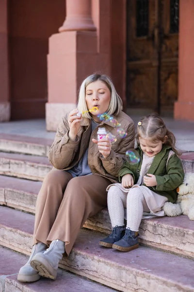 Stylish woman blowing soap bubbles while sitting on street near daughter — Stock Photo
