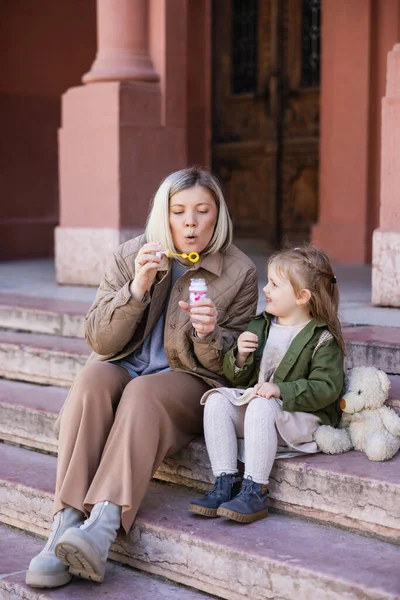 Woman sitting on stairs on street and blowing soap bubbles near daughter — Stock Photo