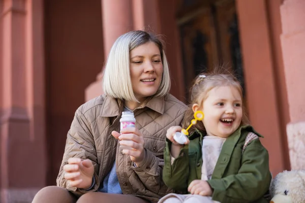 Excited girl with soap bubbles looking away near mom outdoors — Stock Photo