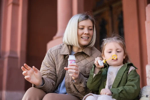 Girl with soap bubbles sitting on urban street near cheerful mother — Stock Photo