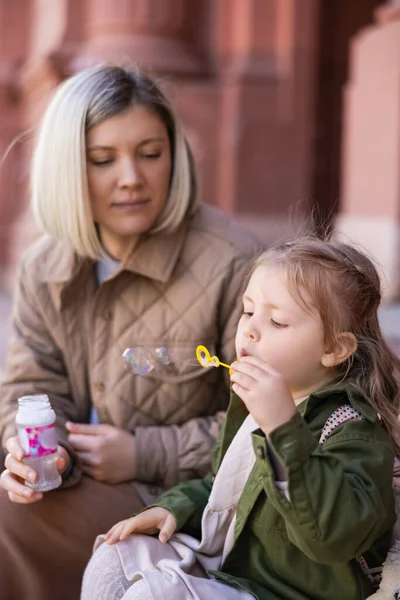 Little girl blowing soap bubbles near blurred mom outdoors — Foto stock