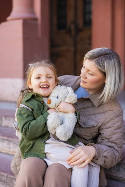 Joyful girl holding soft toy near mom on city street — Stockfoto