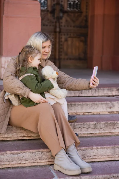 Smiling woman taking selfie on smartphone with daughter and teddy bear on stairs outdoors — Stock Photo