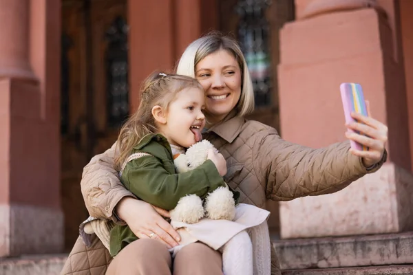 Little girl with soft toy sticking out tongue near smiling mother taking selfie on mobile phone — Stock Photo